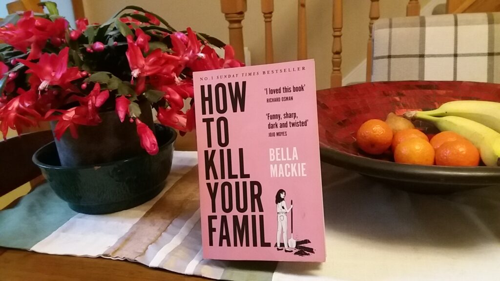 How To Kill Your Family by Bella Mackie propped up on an everyday dining table next to a potted plant and a fruit bowl