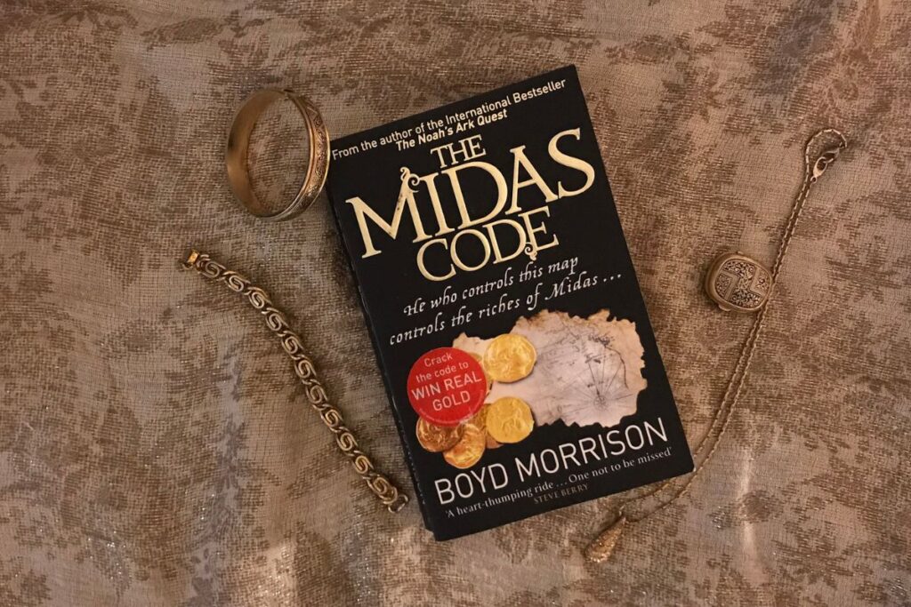 The Midas Code by Boyd Morrison resting on a gold shawl surrounded by gold jewellery