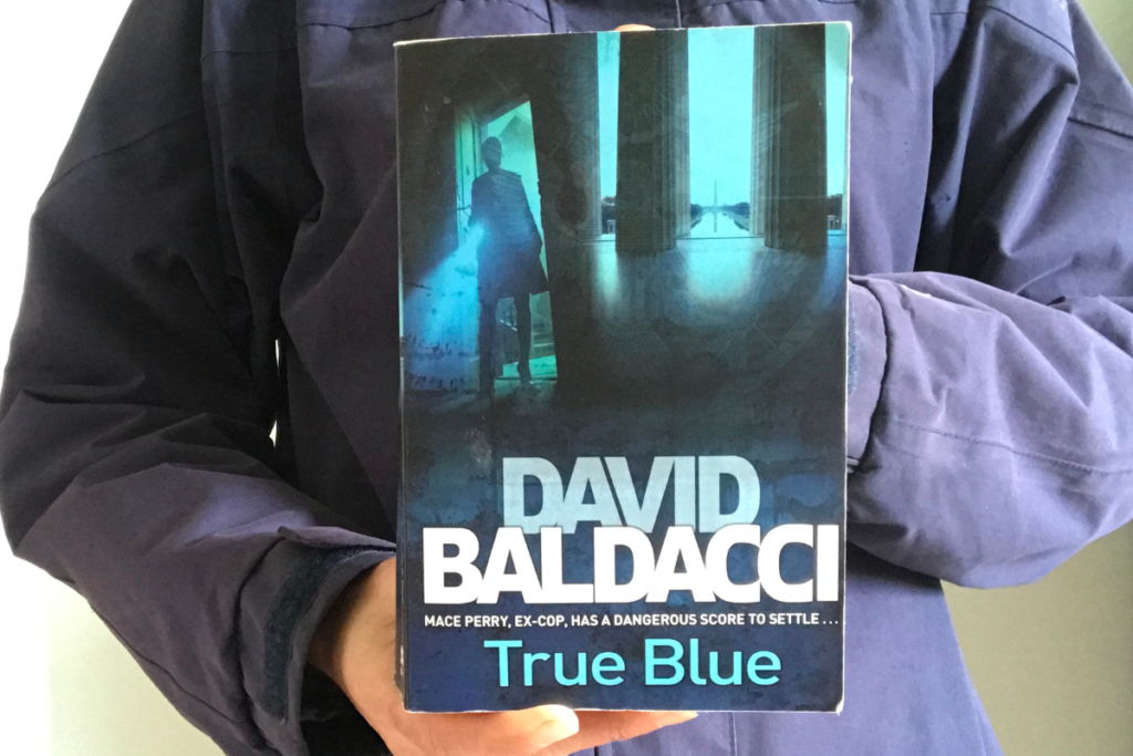 True Blue by David Baldacci held up to the camera by a person in a blue jacket